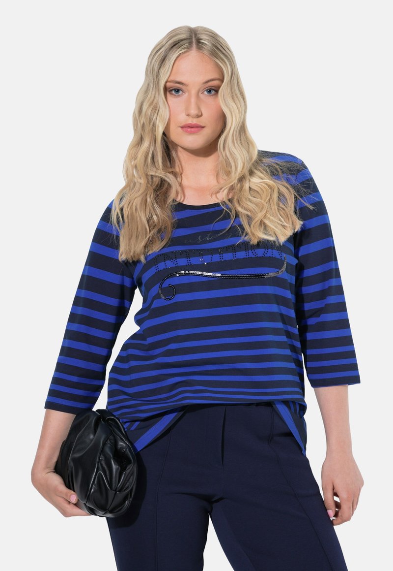 Top a maniche lunghe blu e navy a righe con scollatura rotonda, caratterizzato da dettagli in paillettes sul petto. Modella che tiene una borsa nera.