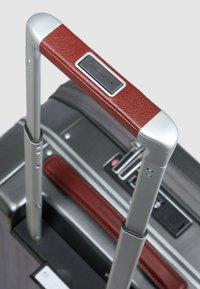 Silberner Koffer mit gebürsteter Textur, ausgestattet mit einem roten Ledergriff, metallischen Beschlägen und einem ausziehbaren Aluminium-Trolley-System.