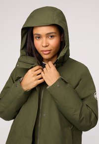 Donkergroene hooded jas met een ritssluiting aan de voorkant, verstelbare manchetten en een logo-patch op de linkerarm. De stof lijkt glad en duurzaam.