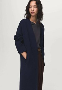 Vrouw in een donkerblauwe lange jas over een grijze top en een bruine broek, staande met één hand in de jaszak tegen een effen achtergrond.