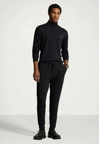 Jeune homme debout, portant une tenue entièrement noire avec un pull à col roulé, un pantalon de jogging, des baskets noires et une montre-bracelet, sur un fond clair et uni.