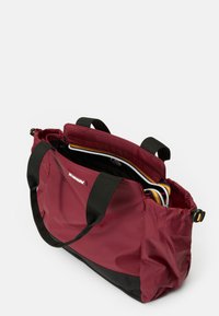 Rode nylon draagtas met zwarte onderkant, voorzien van twee zwarte handgrepen en een geboetseerde bovenkant. Het interieur toont meerkleurige organisatiecompartimenten.