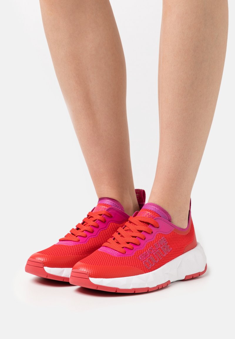Baskets en mesh rouge et rose avec semelles blanches portées aux pieds, avec des lacets orange vif et le logo "Versace Jeans Couture" sur les côtés.