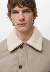 Beige jacka med en mjuk, krämfärgad sherpakrage, med svarta knappar och ett slätt, lätt glänsande tyg.