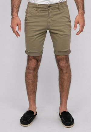 Homme portant un short vert olive retroussé et des chaussures noires sans lacets, debout devant un fond gris uni.