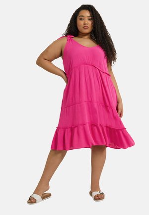 Rosa Sommerkleid mit gestuftem Rüschensaum, verstellbaren Schulterträgern und weichem, strukturiertem Stoff. Getragen mit flachen weißen Sandalen.