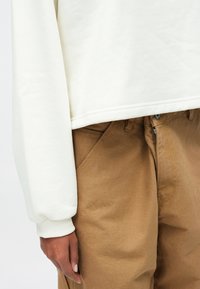 Λευκό, πλεκτό φούτερ με χαλαρή εφαρμογή, με ριμπ τελειώματα στα μανίκια, συνδυασμένο με καφέ, υφασμάτινο παντελόνι με ψηλή μέση.