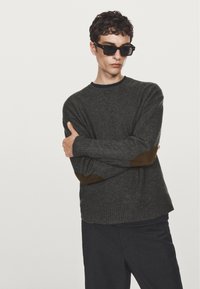 Maglione di lana grigio scuro con toppa marrone sui gomiti, polsini a coste e scollo rotondo, abbinato a pantaloni neri e occhiali da sole.