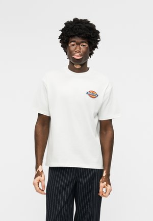 Άτομο με λεύκη φοράει ένα λευκό πουκάμισο με κοντά μανίκια και ένα μικρό πολύχρωμο λογότυπο, καθώς και μαύρο παντελόνι με λεπτές ρίγες, στέκεται μπροστά σε ένα απλό φόντο.