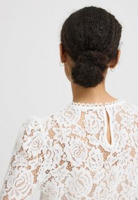 Hvid bluse i spids med blomstermønstre, puffede ærmer og en nøglehulslukning med scallopkant. Gennemsigtig tekstur og delikate designdetaljer.