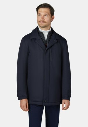 Mann mit Schnurrbart trägt eine dunkelblaue Jacke über einer Reißverschluss-Schicht und einem weißen Rollkragen, steht vor einem schlichten hellen Hintergrund.