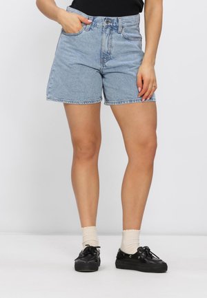 Vrouw draagt lichtblauwe hooggetailleerde spijkershorts, witte enkelsokken en zwarte lage sneakers, staande tegen een effen witte achtergrond.