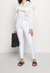 Camisa blanca de botones con un suéter de punto crema por encima, combinada con jeans skinny blancos y sandalias negras. Bolsa negra en mano.