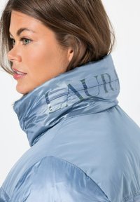 Chaqueta de plumas de color azul claro con un alto cuello. Cuenta con una tela brillante y un sutil estampado "LAURA" en el cuello. Textura suave, cierre con cremallera.