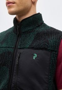 Κοντινή λήψη ενός ατόμου που φοράει ένα σκούρο πράσινο και μαύρο fleece γιλέκο με τσεπάκι στο στήθος με φερμουάρ και ένα μικρό κεντημένο λογότυπο.