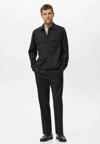 Moški oblečen v črn plašč z žepi na poklopecih, črne hlače in črne čevlje, stoji z rokama v žepih na beli podlagi.