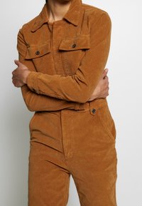 Mono de pana marrón con mangas largas, dos bolsillos en el pecho y un cuello. Presenta detalles de botones y una tela texturizada.