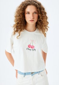 Weißes, kurzärmeliges, verkürztes T-Shirt mit einem Kirschgrafik und dem Text "Cherry Baby" in schwarzer Schrift. Aus weichem Baumwollmaterial.