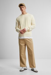 Suéter de punto acanalado amarillo claro superpuesto a una camisa blanca, combinado con pantalones de pierna ancha beige y zapatillas multicolor. Lleva puesta una gorra beige.