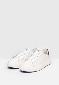 Weiße Ledersneaker mit runder Zehenpartie, flachen Schnürsenkeln, perforierten Seitendetails und einem braunen Lederakzent an der Ferse. Gummisohle mit blauer Umrandung.