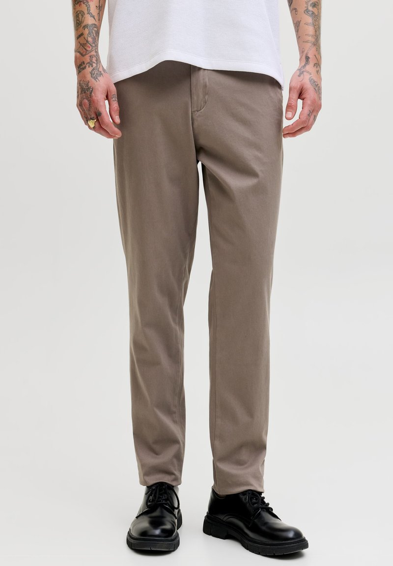 Homme portant un pantalon beige droit, des chaussures noires à lacets, une chemise blanche, et montrant des bras tatoués avec une bague à la main gauche.