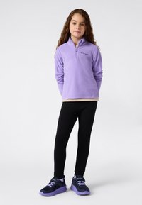 Jeune fille stand avec les mains derrière le dos, portant un pull polaire zippé violet, un legging noir et des baskets noires et violettes sur fond blanc.