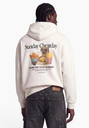 Man with braided hair wearing white hoodie with fast food illustration and text "Sunday Cheatday" on back, paired with black jeans.