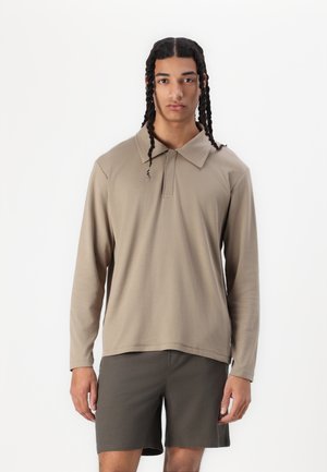 Beige Langarm-Polohemd mit Kragen, das eine lockere Passform und eine weiche Textur aufweist. Kombiniert mit dunkelgrauen Shorts.
