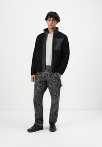 Tânăr îmbrăcat cu o jachetă neagră din polar, o cămașă gri deschis, pantaloni gri cu un model pixelat și adidași negri, stând în fața unui fundal alb.