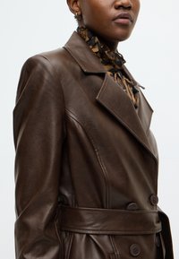 Brun læder dobbeltradet trenchcoat med bælte i taljen, bred krave og tekstureret finish. Har store knapper og skræddersyet pasform.