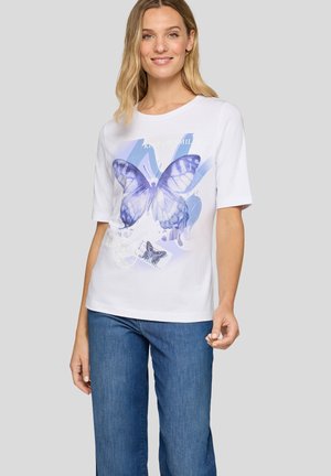 Frau trägt ein weißes T-Shirt mit großem lila Schmetterlingsdruck und hellblauen Jeans vor einem einfarbigen grauen Hintergrund.