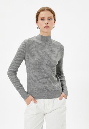 Grauer Strickpullover mit hohem Kragen, der eine gerippte Textur und ein dezentes Diamantmuster aufweist. Das Model posiert mit den Händen in den Taschen und trägt helle Hosen.