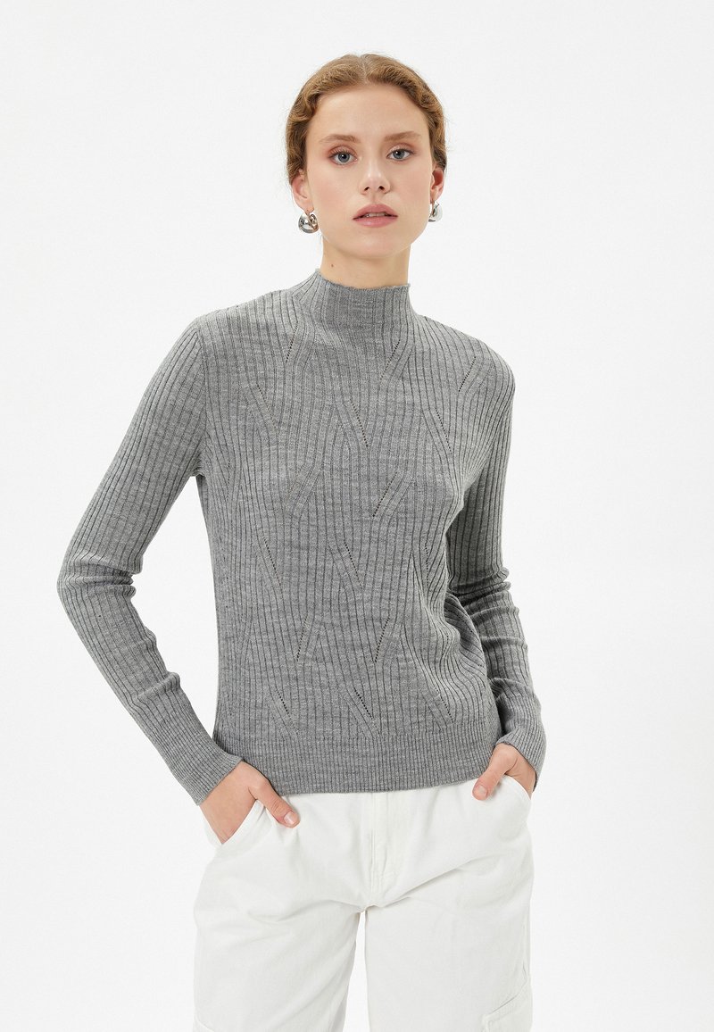 Maglione grigio a maglia con colletto alto, con texture a coste e un sottile motivo a diamante. Il modello posa con le mani nelle tasche, indossando pantaloni leggeri.
