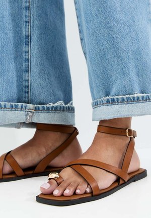 Voeten met bruine leren platte sandalen met gouden accenten, gecombineerd met lichtblauwe opgerolde spijkerbroeken.