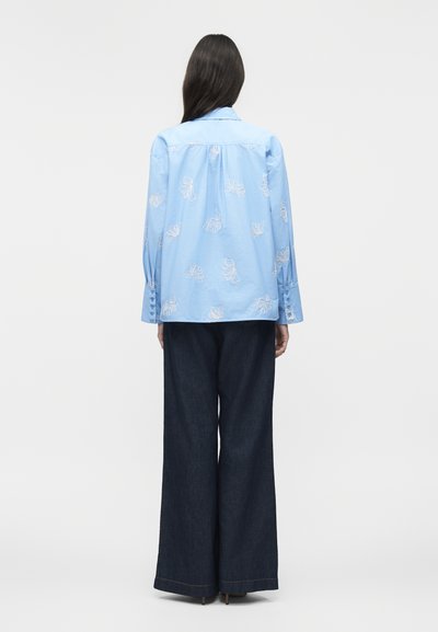Γυναίκα με μακριά σκούρα μαλλιά, που φοράει ένα ανοιχτό μπλε πουκάμισο με κεντητά λουλούδια και φαρδιά σκούρα μπλε παντελόνια, όρθια με την πλάτη στραμμένη.