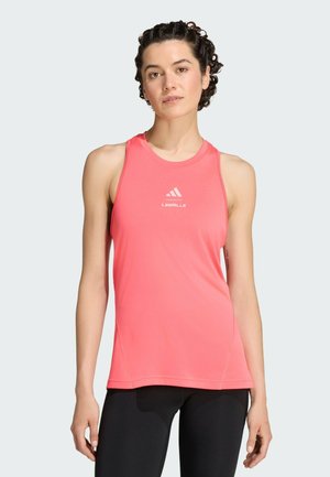 Giovane donna con capelli ricci che indossa un top sportivo Adidas Les Mills rosa acceso senza maniche e leggings neri, in piedi davanti a uno sfondo semplice.