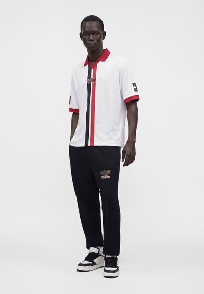Homme portant un polo blanc à rayures verticales rouges et noires, pantalon noir avec logo et baskets noir et blanc, debout devant un fond uni.