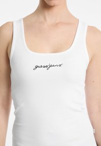 Witte geribbelde tanktop met een ronde halslijn en zwart geborduurd "guessjeans" logo op de voorkant. Zachte textuur en een aansluitend ontwerp.