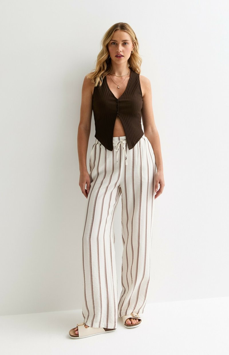 Top sans manches côtelé marron avec des boutons et un col en V, associé à un pantalon large rayé blanc avec des accents marron. Sandales plates montrées.