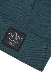 Berretto lavorato a maglia in blu scuro con risvolto, dotato di un'etichetta rettangolare nera con testo bianco, inclusa la marca "SLADE COUTURE."