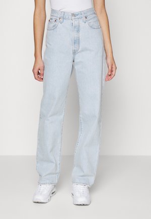 Femme portant un jean droit taille haute bleu clair et des baskets blanches, debout devant un fond clair uni.