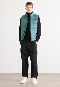 Gilet rembourré teal avec un col haut, fermeture éclair, et un turtleneck noir en dessous ; associé à un jean sombre et des baskets blanches.