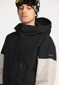 Sort og lysegrå skijakke med høj krave, lynlåslukning og logo detaljer på ærmet. Stoffet har en glat, vandafvisende tekstur.