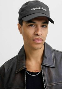 Black cotton cap with "Originals Studio" embroidered in white, worn with a dark leather jacket and ribbed black shirt beneath.
