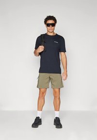 Lyhythihainen tummansininen T-paita, oliivinvihreät cargo-shortsit, mustat lenkkitossut ja aurinkolasit; reppu heitetty yhdelle olalle.