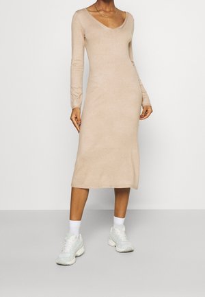 Vrouw draagt een beige midi gebreide jurk met lange mouwen en een V-hals, witte sokken en witte chunky sneakers, staand tegen een effen achtergrond.