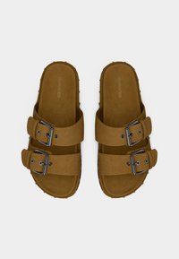 Καφέ σουέτ σανδάλια διαφάνειας με δύο ρυθμιζόμενες μεταλλικές αγκράφες, υφασμένα πέλματα και μικρές λεπτομέρειες με καρφιά κατά μήκος της άκρης.