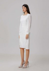 Robe blanche mi-longue avec des manches longues froncées, une fente sur le côté et un décolleté marqué ; accompagnée de talons hauts gris clair. Texture lisse.