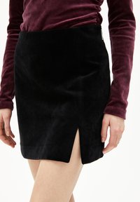 Schwarzer Wildleder-Minirock mit seitlichem Schlitzdetail, strukturiertem Finish und einem klaren, minimalistischen Design. Kombiniert mit einem tiefvioletten Langarmoberteil.