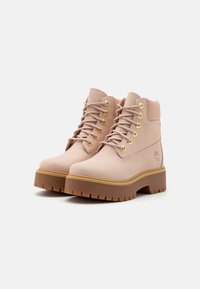 Botas de ante rosa hasta el tobillo con ojales dorados, suela gruesa marrón y cordones texturizados. Diseño sencillo con un logo grabado en el lateral.